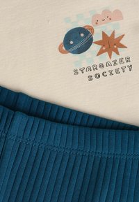 T-shirt in cotone crema con grafiche di un pianeta blu, una stella arancione e nuvole. Tessuto a coste turchese lungo il bordo inferiore, con la scritta "STARGAZER SOCIETY."