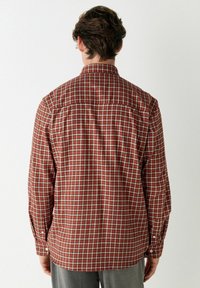 Un homme de dos portant une chemise à manches longues à carreaux rouges et un pantalon gris devant un fond clair.