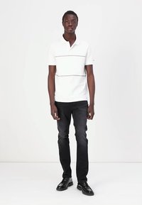 Hvid polo skjorte med en horisontal grå stribe hen over brystet, parret med mørke jeans og sorte sko. Enkel, klassisk design.