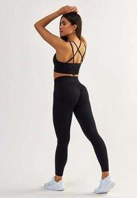 Schwarzer Sport-BH mit überkreuzten Trägern, kombiniert mit hoch taillierten schwarzen Leggings. Modell trägt weiße Turnschuhe. Glattes, dehnbares Material.