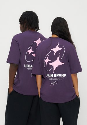 Deux personnes debout dos à la caméra portant des t-shirts violets avec des étoiles pixélisées roses et le texte "Urban Spark" en blanc.