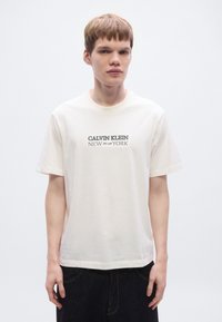 Λευκό βαμβακερό T-shirt με στρογγυλή λαιμόκοψη, κοντά μανίκια και μαύρο σχέδιο στο μπροστινό μέρος. Κανονική εφαρμογή, καθαρές γραμμές και απαλή υφή.