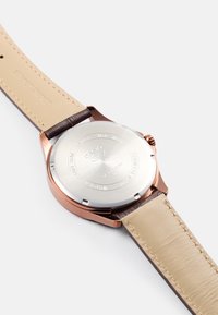 Relógio Casio com fundo em aço inoxidável prateado, caixa em rosé e uma pulseira estreita em pele bege, com costura em tons castanhos.