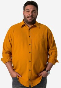 Camisa amarilla mostaza de botón con un bolsillo en el pecho, botones negros y mangas remangadas. Textura suave y diseño casual.