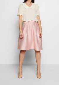 Blusa de color crema con un cuello amplio, combinada con una falda rosa de longitud hasta la rodilla, con pliegues y hecha de una tela suave. Lleva unos tacones altos beige.