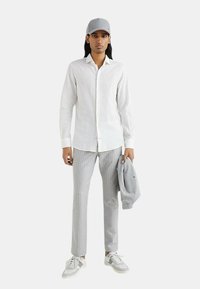 Camicia bianca a maniche lunghe, pantaloni a righe grigie chiaro, berretto grigio, maglione grigio sulle braccia, sneakers bianche e grigie, trame di tessuto morbide.