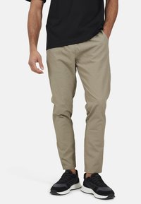 Jasnobeżowe spodnie z teksturowanym wzorem, zwężanym krojem, z elastycznym pasem i bocznymi kieszeniami, w połączeniu z czarnymi butami sportowymi.