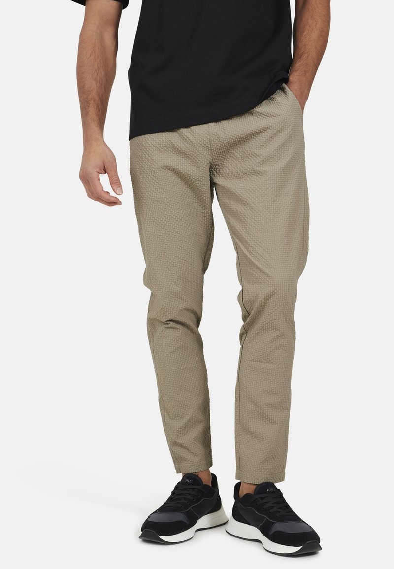 Jasnobeżowe spodnie z teksturowanym wzorem, zwężanym krojem, z elastycznym pasem i bocznymi kieszeniami, w połączeniu z czarnymi butami sportowymi.