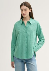 Camisa de botón verde claro con un patrón de puntos texturizados, cuello y mangas largas. Combinada con jeans de mezclilla azul claro.