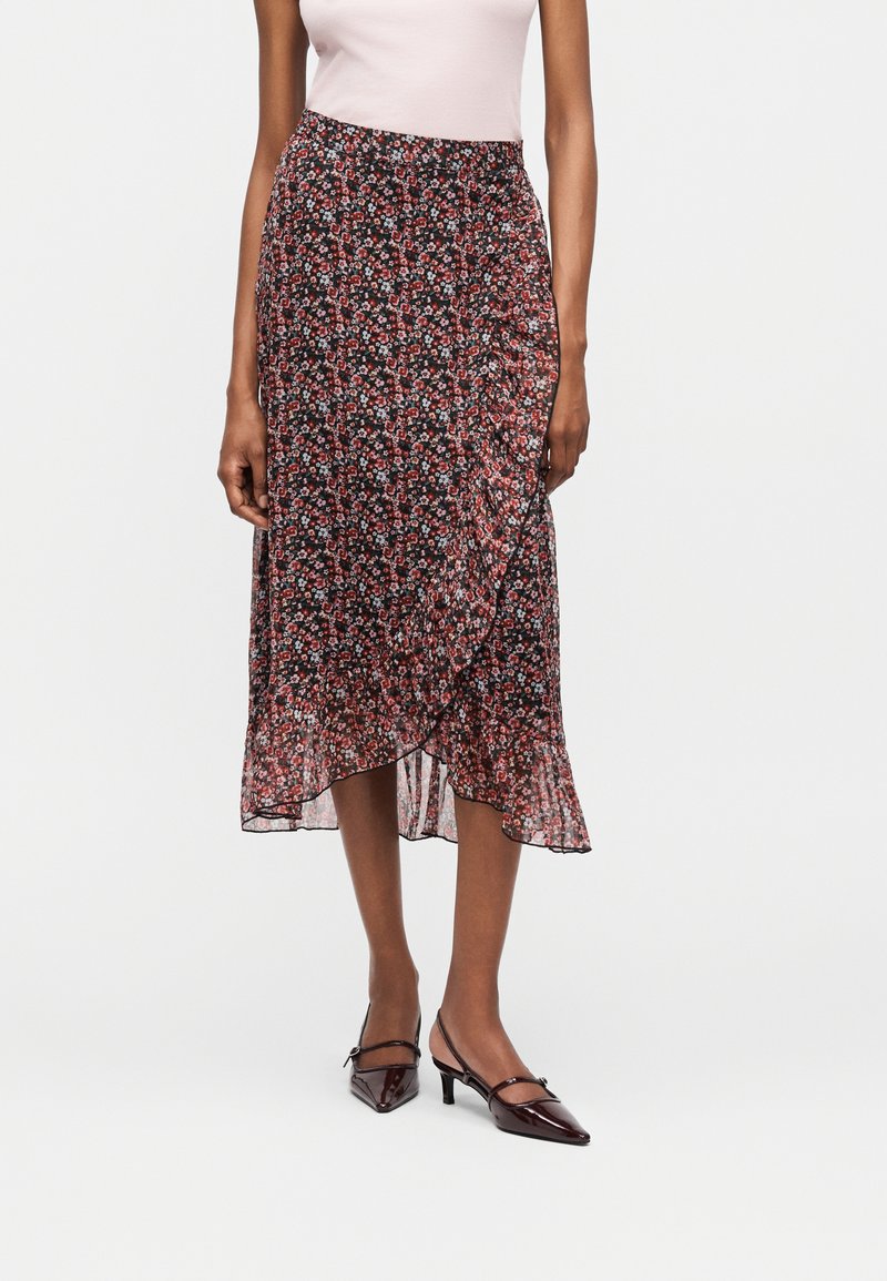 Γυναίκα με μαύρη μεσαία φούστα με μικρά κόκκινα και λευκά λουλούδια, σε συνδυασμό με ανοιχτό ροζ τοπ και σκούρα μυτερά τακούνια.