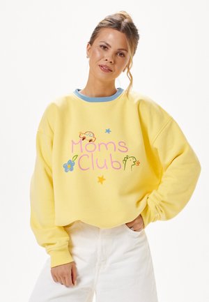 Woman wearing a yellow sweatshirt with blue trim and "Moms Club" text with car, flower, stars, and bird illustrations. White pants, light background.