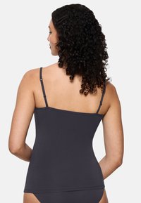 Kobieta z kręconymi ciemnymi włosami widziana z tyłu, ubrana w ciemnoszary top na ramiączkach i pasujące majtki na jasnym tle.