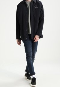 Černá košile na knoflíky přes šedé tričko, kombinována s tmavě modrými džíny a černými teniskami. Na rukou jsou vidět tetování a jednoduchý design.