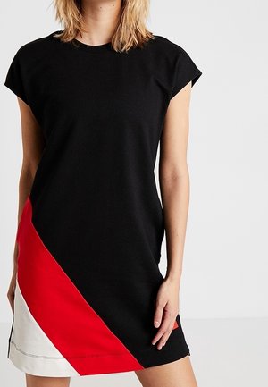 Woman wearing sleeveless black dress with diagonal red and white color block on lower left side, standing against plain white background.