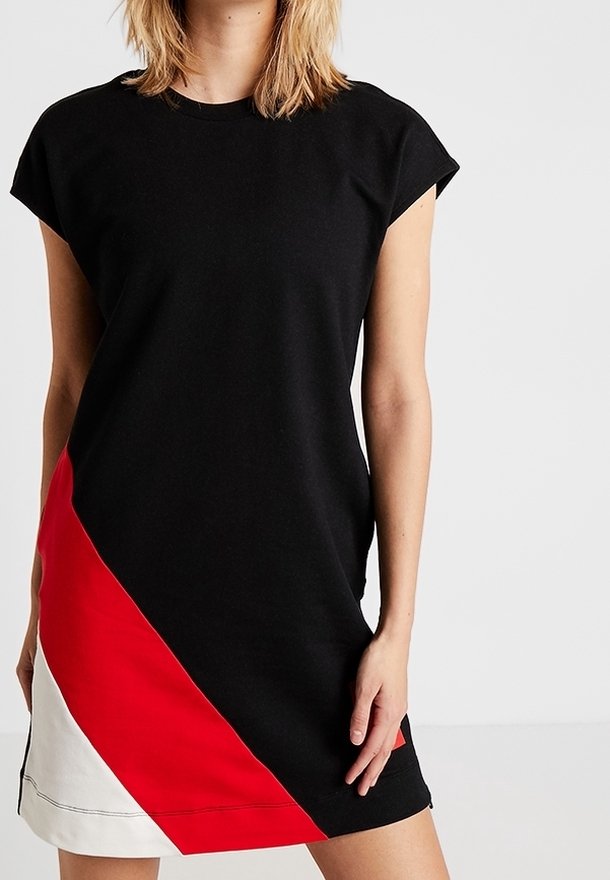 Femme portant une robe noire sans manches avec un bloc de couleur diagonale rouge et blanche sur le côté inférieur gauche, debout devant un fond blanc uni.