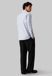 Lyseblå langermet skjorte med knepping-detalj, rett ryggplyndre og svarte dressbukser. Har en avslappet passform og glatt tekstur.