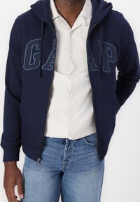 Sweat à capuche zippé bleu marine avec le logo "GAP" en bleu clair contrastant, doté d'une capuche avec cordon de serrage et de deux poches avant. Associé à une chemise claire.