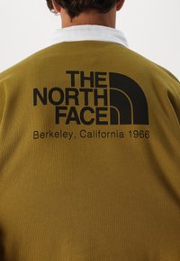 Khaki sweter z białym kołnierzykiem. Czarne logo z napisem "THE NORTH FACE" oraz "Berkeley, California 1966" nadrukowane z tyłu.