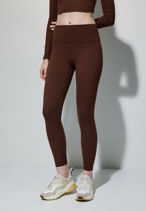 Bruine leggings met een hoge tailleband, een gladde textuur en een strakke pasvorm, gecombineerd met lichtgekleurde sneakers met een gemengde materialenlook.