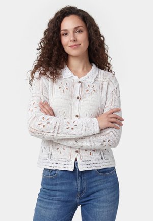 Vrouw met krullend haar, gekleed in een witte gehaakte cardigan met bloemenpatroon en knopen, en een blauwe spijkerbroek, staand met gekruiste armen.
