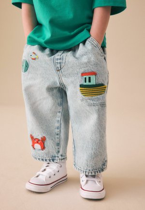 Kind trägt hellblaue Jeans mit gestickten Motiven von Boot, Krabbe, Fisch und Rettungsring, kombiniert mit weißen Turnschuhen und grünem Hemd.