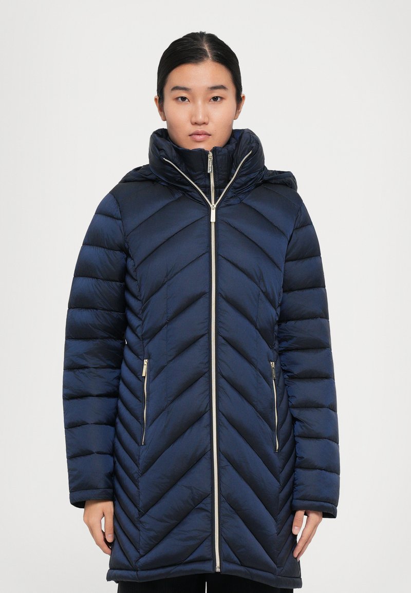 Vrouw in een marineblauwe puffercoat met zigzagpatroon, hoge kraag en ritssluiting aan de voorkant, staand tegen een effen witte achtergrond.