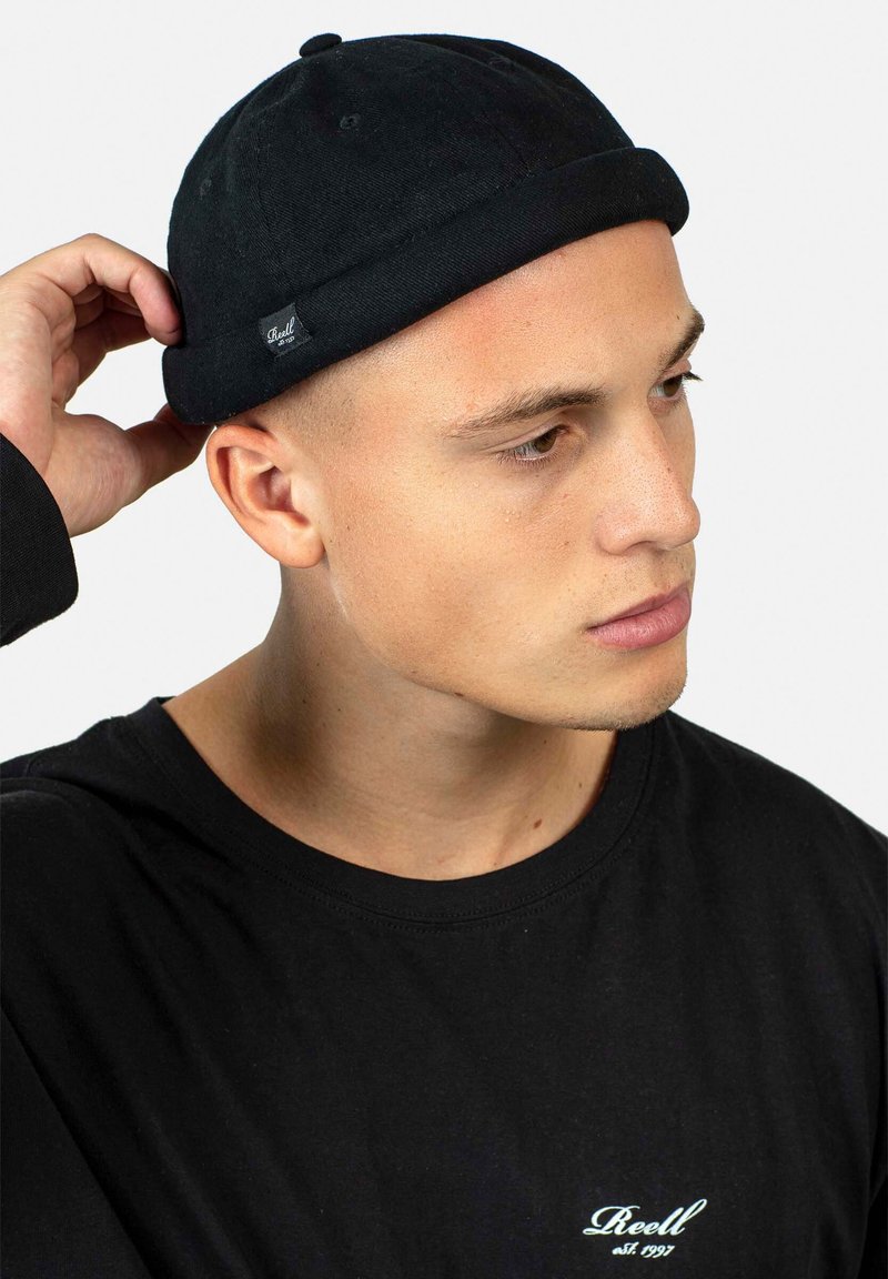 Gorro negro con dobladillo enrollado, hecho de tejido suave. Lleva puesto una persona con una camiseta negra que tiene un pequeño logo en el pecho.