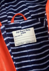 Tissu extérieur orange avec une doublure en polaire rayée douce bleu marine. Étiquette indiquant "JoJo Maman Bébé 12-18 mois", avec un espace pour le nom. Fabriqué en Chine.