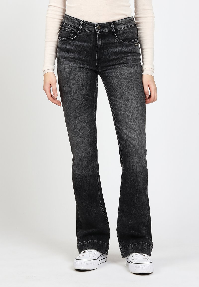 Person, der hochgeschnittene, ausgebleichte schwarze Jeans mit leichtem Schlag und weißen Plateausneakern trägt, steht vor einem schlichten Hintergrund.