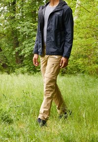 Wasserabweisende schwarze Jacke, graues T-Shirt, beige Cargo-Hose mit Taschen und schwarze Schuhe, getragen in einer grasbewachsenen Außenumgebung.