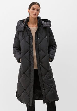 Frau trägt langen schwarzen gesteppter Wintermantel mit Kapuze, beigen Pullover und schwarze Hose, steht mit den Händen in den Taschen vor einfachem Hintergrund.