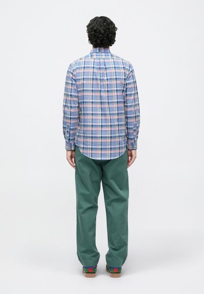 Πουκάμισο με καρό σχέδιο σε μπλε, ροζ και πράσινες αποχρώσεις, με κυρτή μέση, συνδυασμένο με πράσινο παντελόνι. Η πίσω όψη αποκαλύπτει διακοσμητικές ραφές.