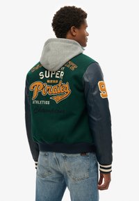 Groene en marineblauwe bomberjack met een grijze capuchon, wollen lijf, leren mouwen en borduursels. Voorzien van gestreepte ribgebreide manchetten en zoom.