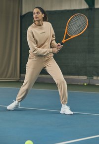 Felpa sportiva beige e pantaloni della tuta con polsini a costine, con logo. Scarpe da ginnastica bianche e una racchetta da tennis sono tenute in una posizione pronta su un campo.