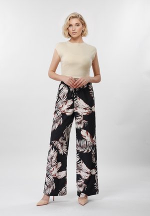 Femme portant un haut beige à manches courtes, un pantalon large noir avec un imprimé de feuilles blanches et brunes, et des chaussures à talons beiges, debout devant un fond blanc.