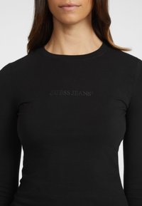 Crna dugorukava uska majica od mekanog materijala. Sadrži reljefni logotip "GUESS JEANS" preko prsa, okrugli izrez i glatku teksturu.