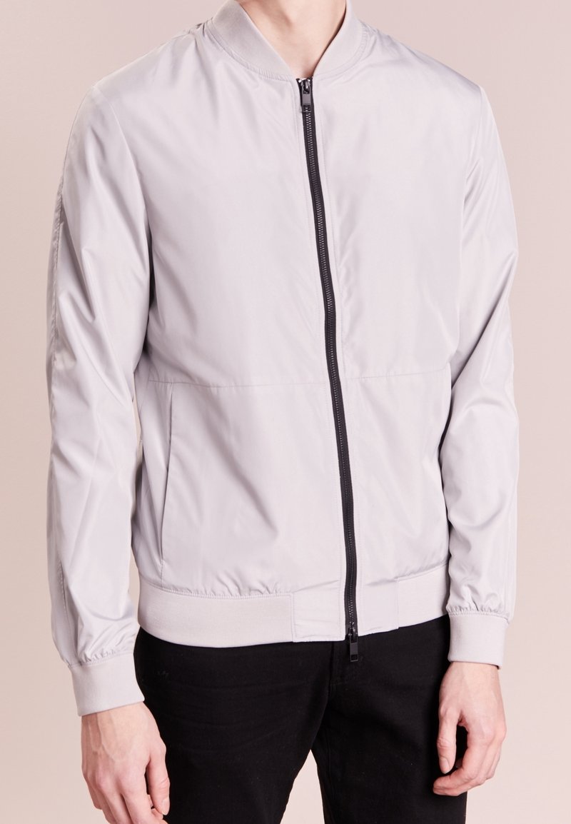 Chaqueta bomber gris claro de tela de nylon, con una cremallera frontal negra, puños acanalados y cinturilla elástica. Textura suave con un diseño minimalista.