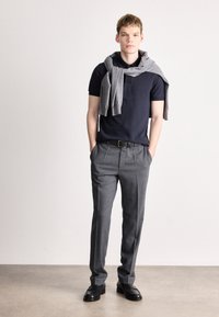 Ναυτικός πόλο πουκαμίσα, γκρι ζακέτα ριγμένη στους ώμους, γκρι πλισέ παντελόνι, μαύρη ζώνη και παπούτσια, σε απλό φόντο.