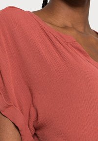 Blusa color ruggine in tessuto texture, con scollatura a V e maniche corte. Presenta pieghe sulle cuciture delle spalle.