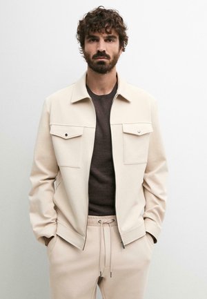 Mand med krøllet hår og skæg iført en beige jakke over en mørkebrun skjorte og beige bukser med snøre, står mod en hvid baggrund.