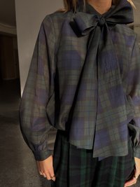 Translucent plaid blouse in navy and green, featuring a large bow at the collar and billowed sleeves. The fabric has a lightweight texture.