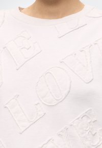 Gros plan d'un sweat-shirt blanc avec des lettres en relief et texturées formant le mot « LOVE » qui se chevauchent.