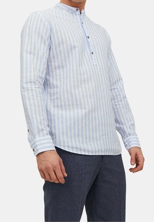 Mężczyzna w jasnobłękitnej i białej koszuli w pionowe paski z długim rękawem i zapinanym kołnierzem oraz ciemnoniebieskich spodniach na jednolitym tle.