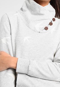 Sudadera gris con cuello alto, tres botones marrones en el cuello y un patrón de aves. La tela es suave con una textura lisa.