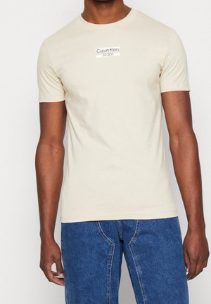 Man draagt een beige Calvin Klein Jeans T-shirt met korte mouwen en blauwe spijkerbroek, staand met de armen ontspannen langs het lichaam.