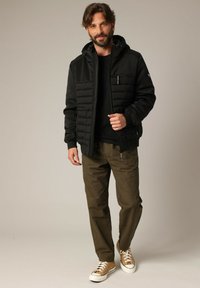 Homme portant une veste à capuche noire, une chemise noire, un pantalon olive et des baskets beige se tenant devant un fond neutre.