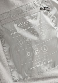 Grått inneretikett på ett ytterplagg med produktinformation, skötselsymboler och kvalitetsattribut som vattentät och vindtät.