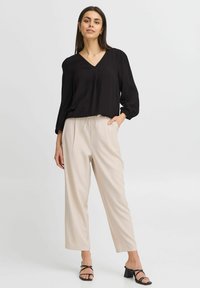 Blusa nera a V scollatura ampia in tessuto testurizzato, abbinata a pantaloni color crema a vita alta. Modella che indossa tacchi neri con cinturini.