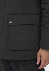 Parka negra con gran bolsillo frontal con cierre a presión y una tela texturizada. Los extremos de las mangas están terminados con un dobladillo limpio.