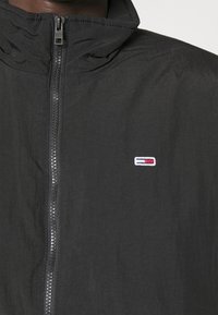 Sort zip-jakke i let stof med høj krave og et lille broderet logo på brystet. Tekstureret overflade med glans.